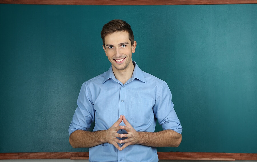 Junger Lehrer steht vor einer leeren Tafel und lächelt in die Kamera